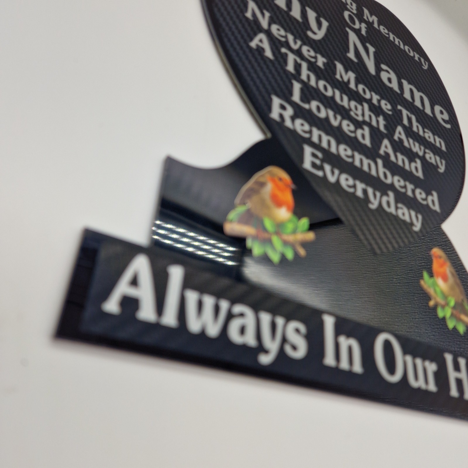Angled close view of silver lettering and robin illustrations on a black heart-shaped memorial plaque.